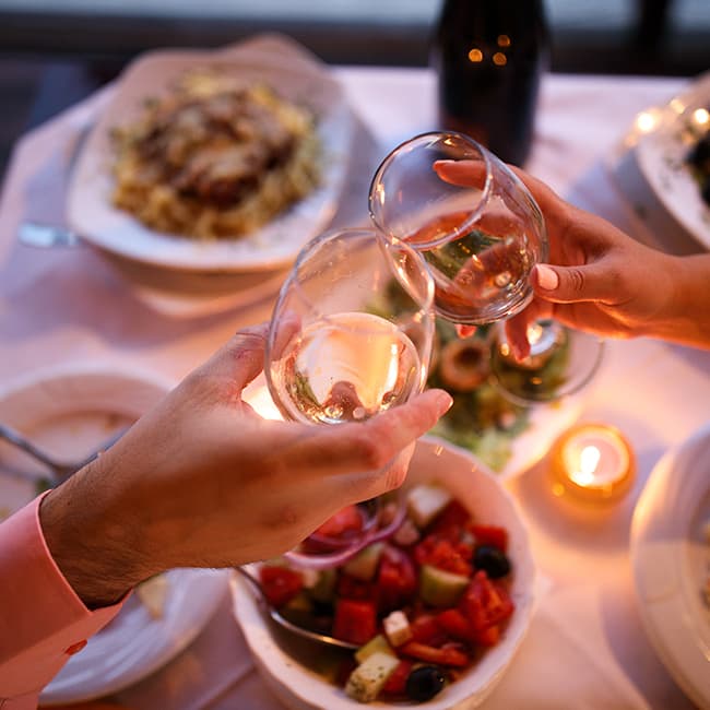 Two adults touching wine glasses over a spread of food in a romantic setting